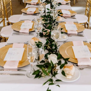 Fancy table setting with gold chargers and wineglasses with greens along center.