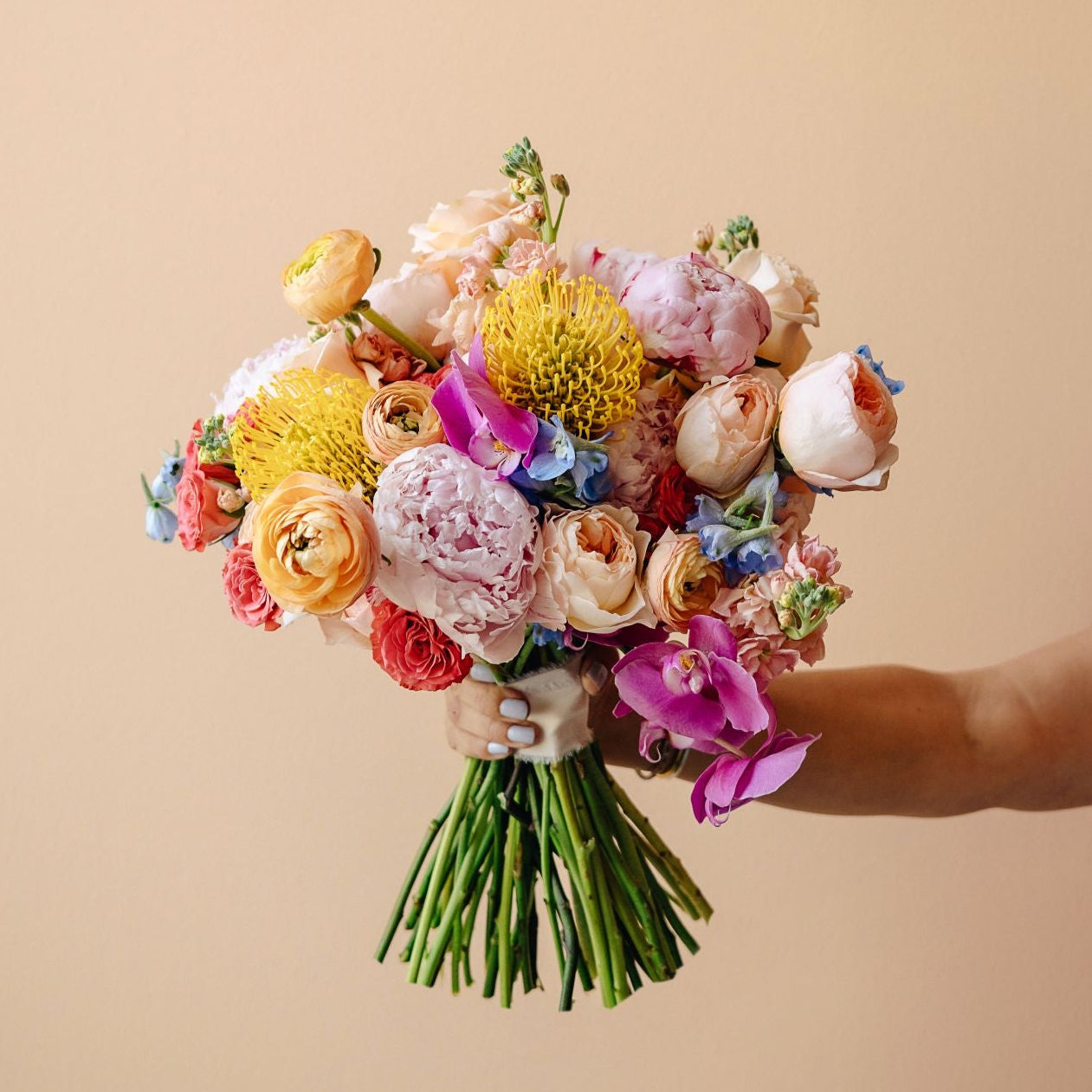 A hand holding a vibrant, multicolored bouquet of dried and fresh flowers against a solid beige background.