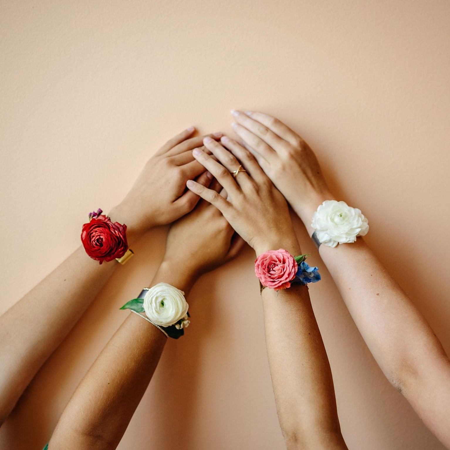 Four hands stacked together wearing four different fabric flower wrist corsages in red, white, pink, and white