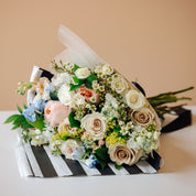 A mixed floral bouquet with fresh white, blush pink, and cream roses, green foliage, and small blue flowers