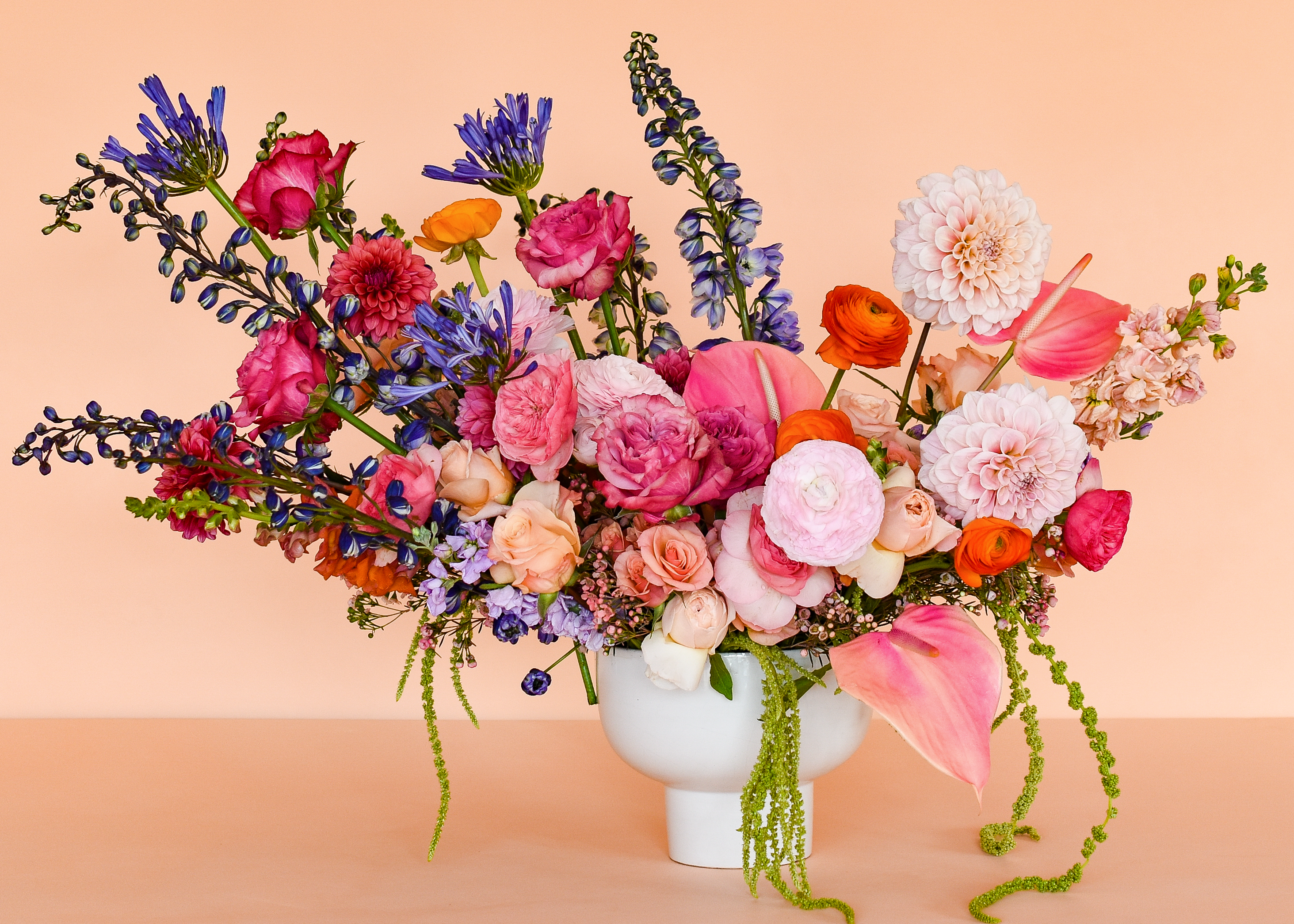 A vibrant floral arrangement featuring pink, red, purple, orange, and white flowers in a white ceramic vase, set against a solid peach background.