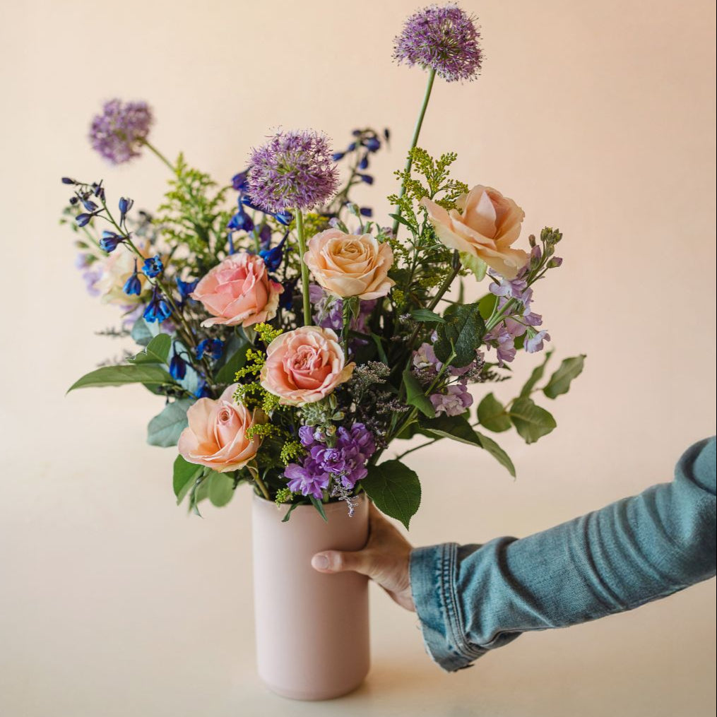 Pale peach roses, bright purple allium, vibrant blue delphinium and more arranged in a vase