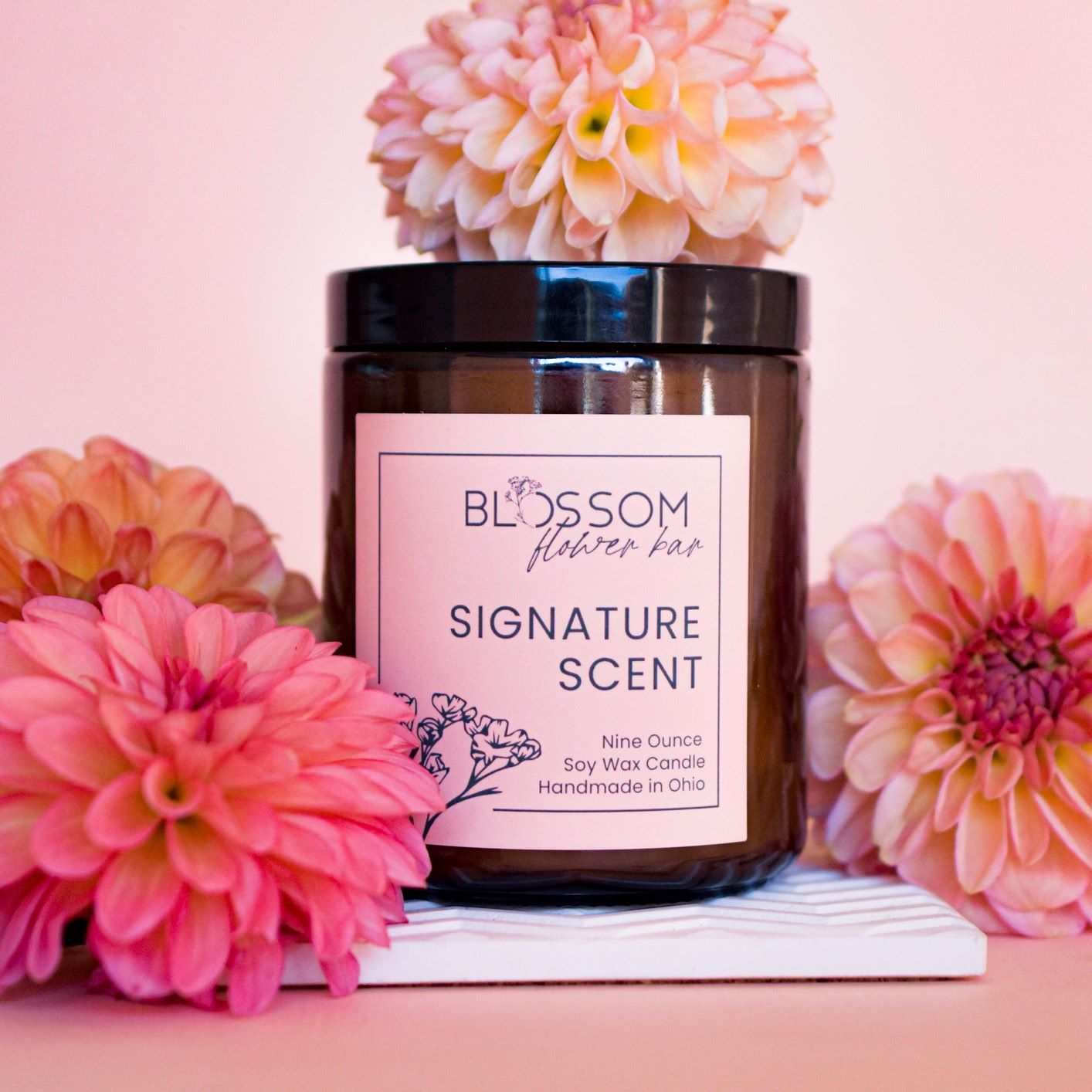 An amber glass candle with Blossom&#39;s label for Signature Scent. The candle is surrounded by peach colored dahlias. 