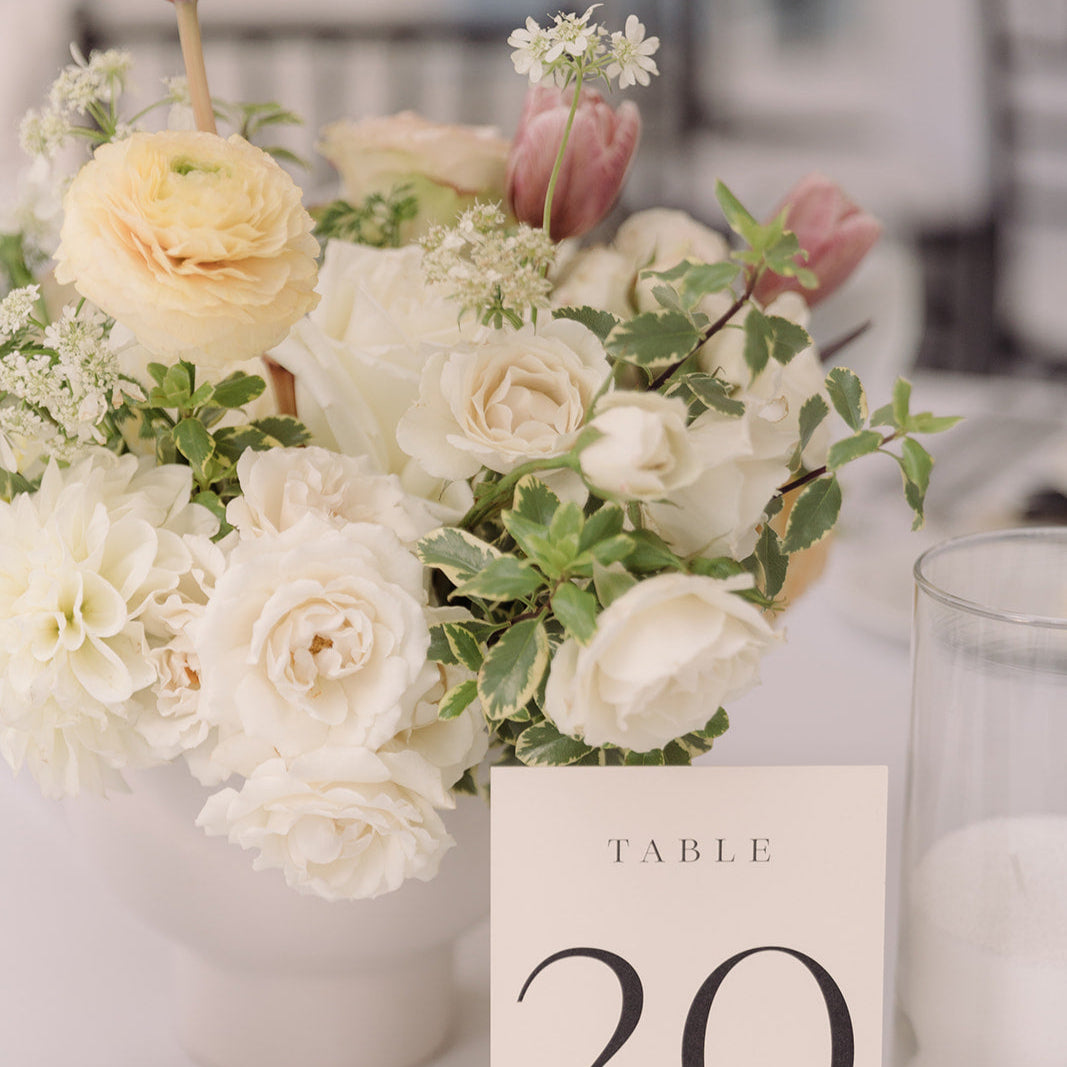 a vased arrangement with peach ranunculus, pink tulips, white spray roses complements a table number and candles